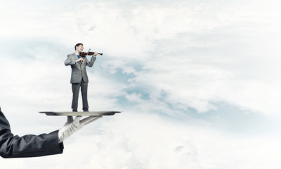 Businessman on metal tray playing violin against blue sky background