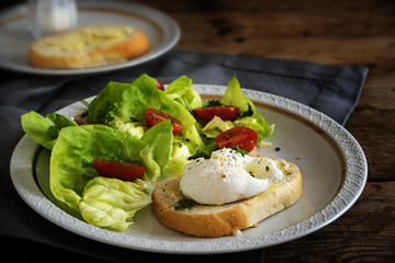 Poached egg on baguette slice with salad and tomatoes, white plate on a dark rustic wooden table, delicious breakfast or snack