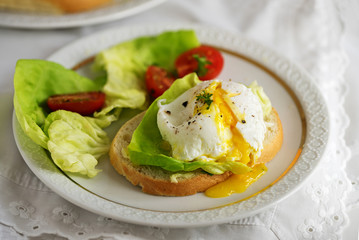 Poached egg with running yolk on baguette slice with salad and tomatoes on a white plate, delicious breakfast or snack