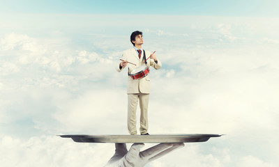 Young businessman on metal tray playing drums against blue sky background