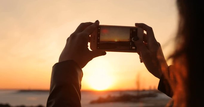 A woman is taking pictures of a sunset on a smartphone