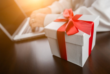 White Gift Box with Red Ribbon and Bow Near Man Typing on Laptop Computer.