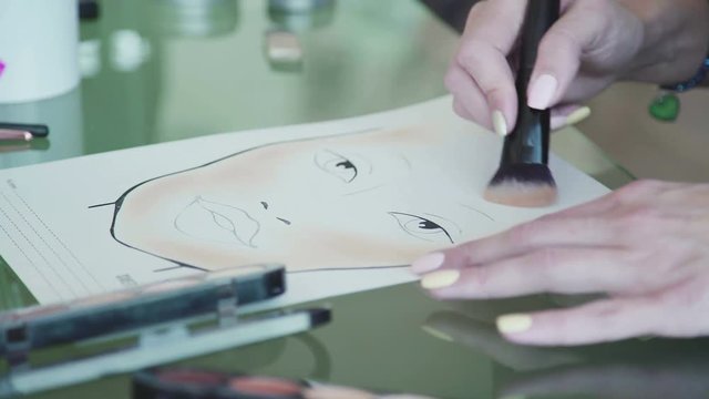 Close-up: make-up artist puts the tone on the face chart using brush. Make-up artist creates a make-up sketch on the face chart. Schematic image of a face on watercolor paper and applies make-up.