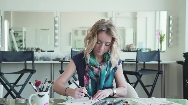 Make-up artist creates a make-up sketch on the face chart. Woman in a draws a facechart. Stylish blonde woman in a bright studio sits at a table and draws a face chart using makeup and brushes