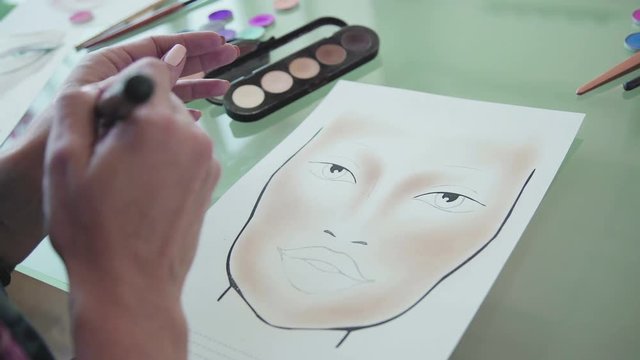 Make-up artist creates a make-up sketch on the face chart. Inspiration: make-up artist draws face-lines on watercolor paper, using professional cosmetics. Face sculpting - training on the face-chart.