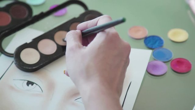 Young woman in a bright studio draws a facechart. The make-up artist draws a schematic image of a face on watercolor paper and applies make-up. Make-up artist creates make-up sketch on the face chart