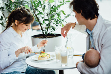 Fototapeta na wymiar Young happy family with newborn baby boy having breakfast in cafeteria. Dad holding little son while mom eating her breakfast. People, healthy lifestyle, family and food concept