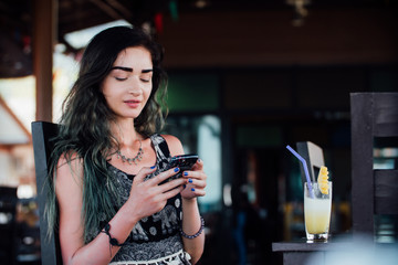The girl typing in the phone, sitting in a cafe with fruit pineapple juice in a glass and a tube.