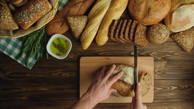 Male hands cutting whole grain bread on the wooden table. Different types of bread. Top view. Slow motion