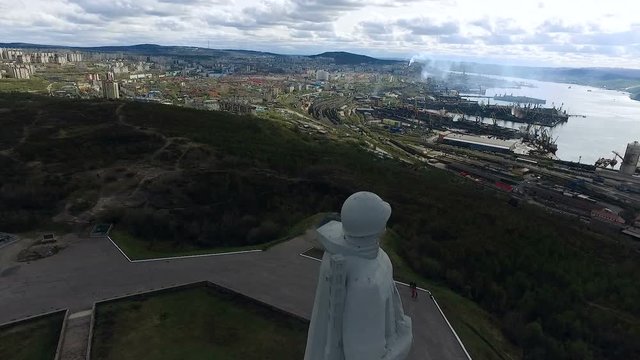 Tall Soviet Russian Soldier Monument on Hill. River Bank Port. Mountains Around City. Smoke Raising From Dock Station. Cloudy Summer Sky. Top View