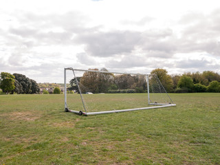 Obraz na płótnie Canvas An Empty and Unused Goal Post with A White Net in the Middle of A Park with Grass and Soil on the Ground, Wheels on the Frame to Help Move it and Houses and Trees in the Background, Used For Football