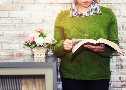 woman is  reading bible in front of white brick wall near book cabinet