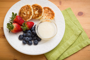 Top view on white plate with cakes made of cottage cheese with blueberries, strawberries and sweet sause on wooden table. Healthy snack. Breakfast idea.