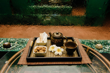 Traditional Thai famous ceremony bronze antique tea-kettle on wicker salver with lotus flowers, cup, sugar and cookies on rattan table with glassy surface with abstract waterfall wall on background.