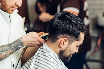 Creating new hair look. Young bearded man getting haircut by hairdresser while sitting in chair at...