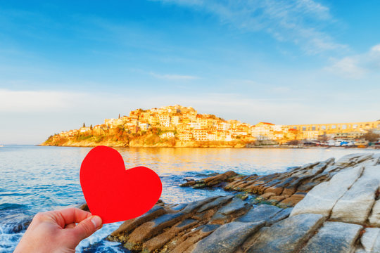 I love Greece concept. Person keep paper red heart in hand at greek medieval summer resort city Kavala. Sunrise scenery.