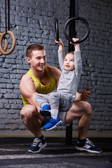 Happy father and little son exercising with gimnastic rings and smiling against brick wall in the gym.