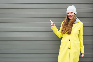 Long-haired stylish girl in a yellow coat and hat smiles and points with her finger to the side.