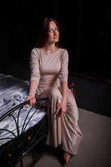 Fototapeta na wymiar Beautiful yong woman sitting on the bed indoors. Wore long classic dress and shoes. Low key studio shot