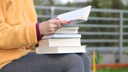 Kobieta w żółtej kurtce i szarych getrach siedzi w parku, na kolanach ma stos książek, w ręku...