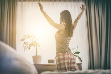 Young woman stretches out in the morning after waking up looking out the window.