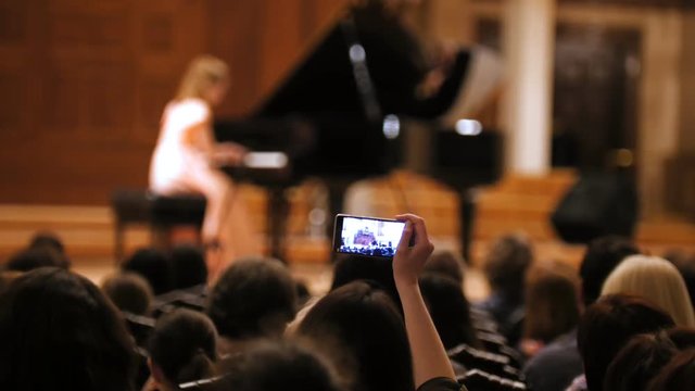 Audience in concert hall during performing piano girl- people shooting performance on smartphone, music opera