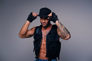 Tattooed male dressed in a jeans, cap and waistcoat on a naked torso.
