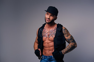 Tattooed male dressed in a jeans, cap and waistcoat on a naked torso.