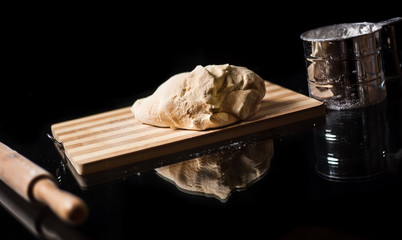 Dough on a wooden board, a rolling pin and a glass for sifting flour are reflected in a glass table