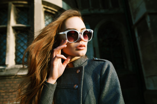 Outdoor close up portrait of young beautiful woman with long hair wearing stylish light blue sunglasses, grey coat posing on street. Model looking aside, touching her face. Female fashion concept
