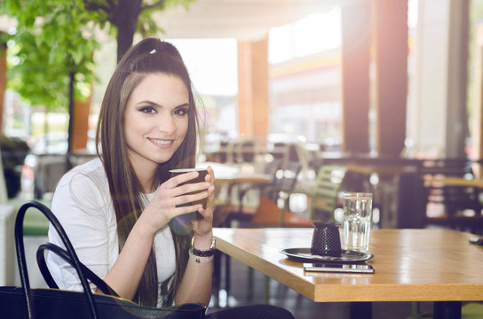 young woman sitting in a cafe, drinking coffee, relax after work and looking at the camera with a smile