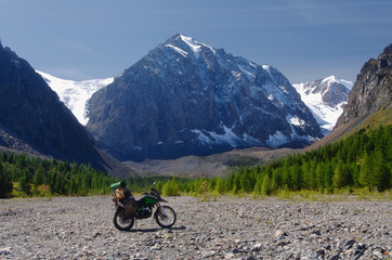 Fototapeta na wymiar Motorcycle traveler with suitcases standing on a stones in high mountain valley among the snow rocks with a glacier surrounded by larch trees under the blue sky Aktru Altai Mountains Siberia Russia