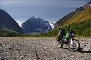 Motorcycle traveler with suitcases standing on a stones in high mountain valley among the snow rocks with a glacier surrounded by larch trees under the blue sky Aktru Altai Mountains Siberia Russia