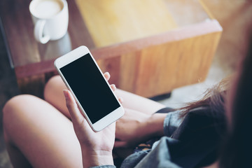 Mockup image of a woman holding white mobile phone with blank black screen with coffee mug on wooden table in cafe