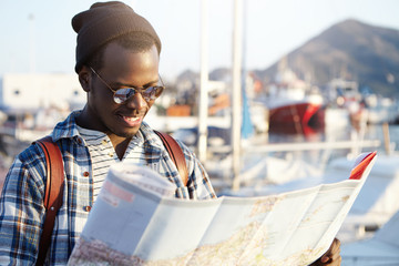 Side view of dark-skinned tourist with backpack in trendy hat and sunglasses examining directions using his paper road map on blurred background of yacht park or club in picturesque resort town