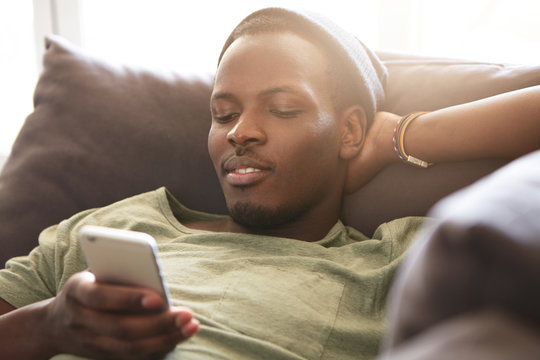 Indoor Shot Of Smiling Black Young Man Surfing Internet On Mobile Phone, Messaging Friends Online And Checking Newsfeed Via Social Media While Relaxing At Home, Lying On Couch With Hands Behind Head