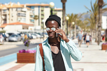 Outdoor shot of handsome dark-skinned fashionable man in trendy headwear and sunglasses talking on mobile phone, walking around metropolis, stopped as he sees beautiful woman in front of him
