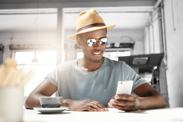 Handsome young Afro American male tourist wearing hat and trendy shades smiling happily while reading sms on smart phone, messaging his girlfriend online using free wi-fi, having coffee at cafe