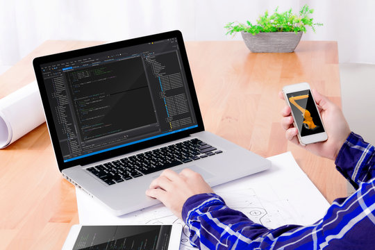 Software engineer's workplace. Engineer's hands working on laptop and holding smartphone with kuka on display. Note: Program code was got from github with free license for commercial use (MIT)