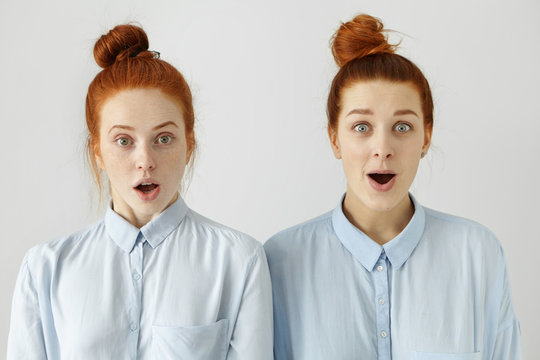Studio shot of two sisters or friends looking alike with their identical blue shirts and same hairstyles looking at camera in full disbelief, shocked or surprised with some news, gossips or rumours
