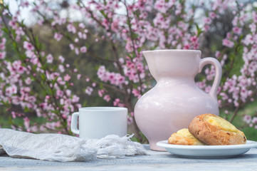 Porcelain pink jug, white cup and bakery on a table of white boards against the background of a flowering bush