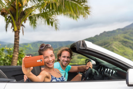 Car holiday selfie. Couple having fun on summer vacation road trip taking smartphone pictures during travel. Multiracial young people driving convertible.