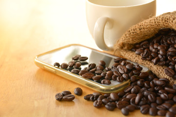 Coffee beans in the sack put on the smart phone with cofee cup on wooden table.