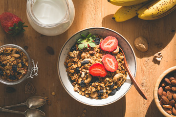 Granola, milk and fruits. Tasty and healthy breakfast: bowl of granola with fresh strawberries, jug of milk, almonds, bananas and spoons on wooden table with ray of morning sunshine. Top view