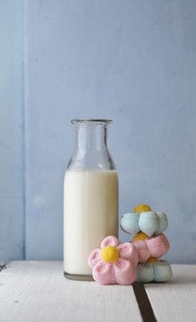 A child's bottle of milk with a sweet treat of flower shaped marshmallow treats in pink and blue on white wooden table , blue background