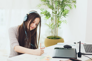 Teenage girl graphic designer using notebook and graphic tablet with stylus while studying at home