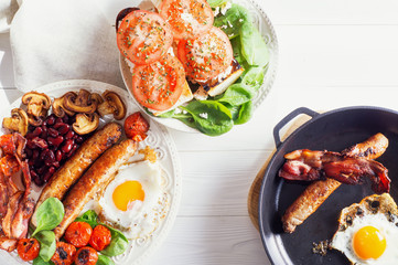 Delicious full English breakfast with fried egg, bacon, sausage, tomatoes, beans and mushrooms.  Traditional english breakfast on white wooden background, top view, flat lay.