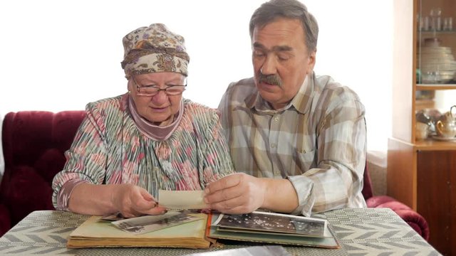 An elderly couple man and woman are looking at their old photos at home and talking. A man with a mustache, a wife with glasses
