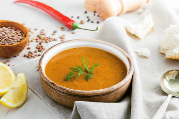 Healthy vegetarian lentil soup in a rustic clay bowl , piece of bread and lemon slices on a white wooden table. Delicious healthy meal served on a canvas tablecloth.