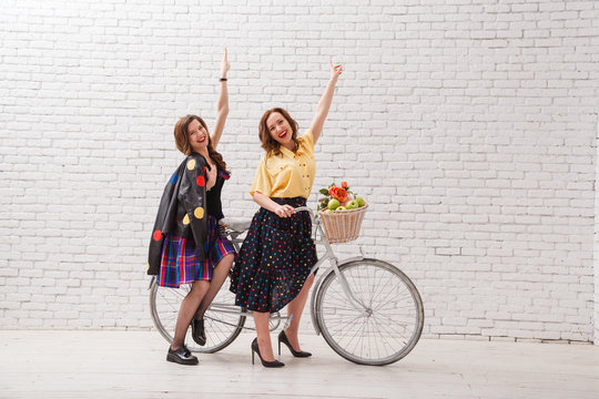 Two Happy Women In Summer Dresses Are Riding Together On A Retro Bike And Gesture Hands Forward.